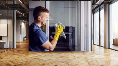 Young bearded smiling positive man with rubber gloves cleaning window with cloth. After rain there is always stains on glass. Wall mural