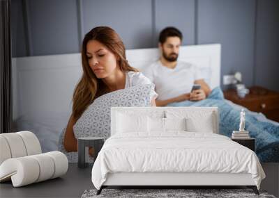 Young beautiful gorgeous sad girl sitting and hugging pillow on the bed in front of her boyfriend with mobile in hands. Wall mural