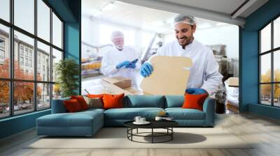 Young Caucasian employee in sterile uniform packing goods in boxes. In background supervisor holding tablet and counting boxes. Wall mural