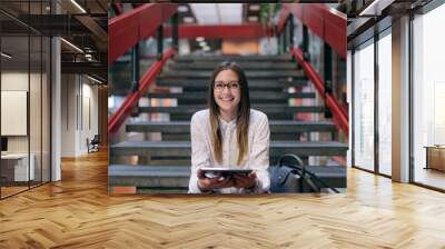 Young Caucasian female student with brown hair and eyeglasses using tablet while sitting on the stairs in college building. Next to her bag. Wall mural