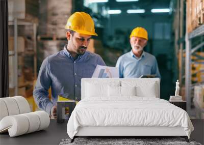 Young Caucasian man with serious face wearing helmet and looking at chart while standing in warehouse. Wall mural