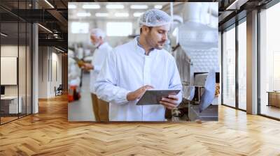 Young Caucasian serious supervisor evaluating quality of food in food plant while holding tablet. Man is dressed in white uniform and having hair net. Wall mural