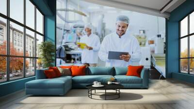 Young Caucasian smiling supervisor in sterile white uniform using tablet while standing in food plant. In background older one worker controlling machine. Wall mural