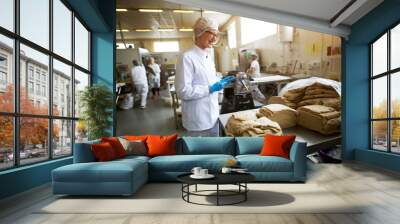 Young cheerful female worker in sterile cloths holding a tablet and checking the quality of cookie dough mixture before further processing. Wall mural