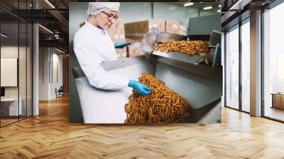 Young female worker in sterile clothes is taking a sample of salt snacks from production line. Wall mural