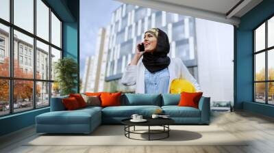 Young positive attractive stylish female muslim architect standing outdoors with helmet under armpit and having business call. Wall mural