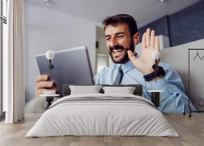 Young smiling bearded handsome businessman sitting at home in char, having conference call and waving. Wall mural