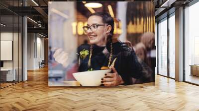 Young smiling girl sitting in restaurant and enjoying her salad for lunch. healthy lifestyle concept. Picture taken from outside. Wall mural