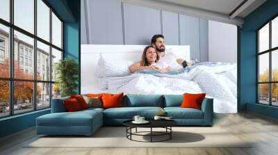 Young smiling happy attractive Caucasian couple lying in bed and watching television in the morning. Bedroom interior. Wall mural