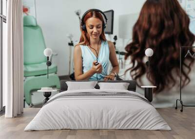 Audiologist doing impedance audiometry or diagnosis of hearing impairment. An beautiful redhead adult woman getting an auditory test at a hearing clinic. Wall mural