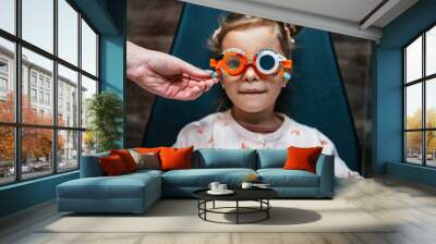 Beautiful and adorable little girl with optometry glasses receiving ophthalmology treatment. Doctor ophthalmologist checking her eyesight with modern equipment. Focus on glasses. Wall mural