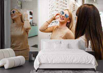 Beautiful and fashionable woman choosing eyeglasses frame in modern optical store. Female seller specialist helps her to make right decision. Wall mural