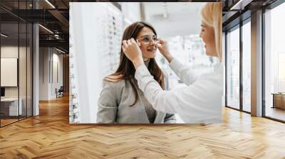 Beautiful and fashionable woman choosing eyeglasses frame in modern optical store. Female seller specialist helps her to make right decision. Wall mural