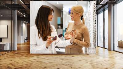 Beautiful and fashionable woman choosing eyeglasses frame in modern optical store. Female seller specialist helps her to make right decision. Wall mural