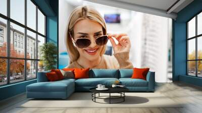 Beautiful and fashionable woman choosing eyeglasses frame in modern optical store. Wall mural