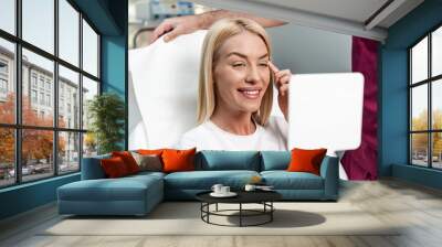 Beautiful and happy young woman sitting in medical chair and looking in the mirror. She is satisfied after successful beauty treatment with hyaluronic acid fillers or botulinum toxin injections. Wall mural