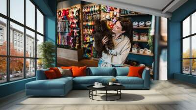 Beautiful young woman enjoying in modern pet shop together with her adorable brown toy poodle. Wall mural