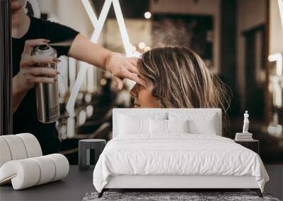 Beautiful young woman with long curly hair in hair salon. Professional hairdresser styling with hairspray. Wall mural