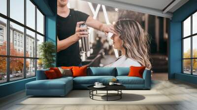 Beautiful young woman with long curly hair in hair salon. Professional hairdresser styling with hairspray. Wall mural