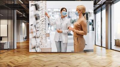 Blonde fashionable woman choosing eyeglasses frame in modern optical store. Female seller specialist helps her to make right decision. They are wearing protective face mask against virus pandemic. Wall mural