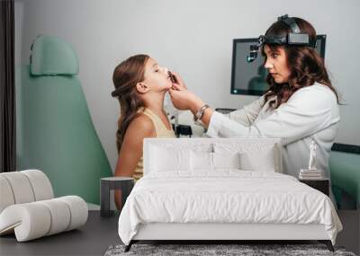 Female audiologist examining girl ear using otoscope in doctors office. Child receiving a ear exam. Nose and throat medical examination. Healthcare and medicine concept. Wall mural