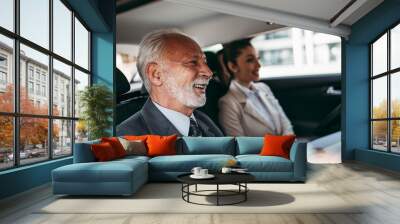 Good looking senior business man and his young woman colleague or coworker sitting on backseat in luxury car. Transportation in corporate business concept. Wall mural