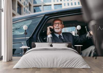 Good looking senior business man sitting on backseat in luxury car. He opens car doors and going or stepping out. Big business building in background. Transportation in corporate business concept. Wall mural