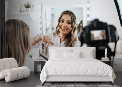 Happy young woman streaming a beauty makeup vlog from home or workshop. Beautiful online content creator cosmetician applying makeup and explaining some work tools. Vlogging and online channel work. Wall mural