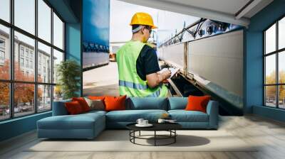 Professional male industrial truck driver with yellow protective helmet performs technical inspection of the vehicle as safety measures before next drive. Wall mural