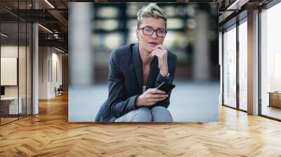 Young happy business woman sitting in front of big modern building. She holding smart phone and seriously looking in distance. Wall mural