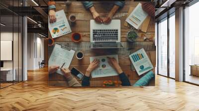 Top view of an office table with multiple hands working on laptops, desks and coffee mugs scattered around it, all holding different types of documents or charts in their hand. Wall mural