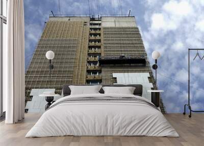 Bottom up view of being renovate high building with insulation on facade and construction cradle under sky with soft white clouds Wall mural