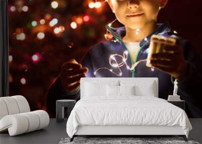 Cute boy playing with Christmas lights and Christmas tree in the background. Wall mural