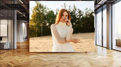 Redhead woman in white summer dress standing on sand in nature at sunset and looking at camera  Wall mural