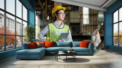 Asian woman engineer industry heavy worker wearing hardhat and holding tablet standing at machine area in factory, engineering industrial concept. Wall mural