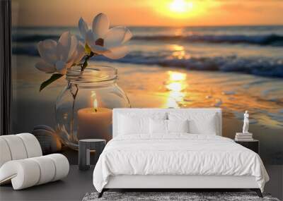 A lit candle in a glass jar with white flowers sits on a sandy beach at sunset. Wall mural