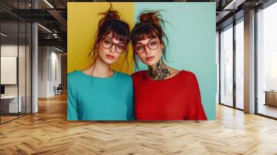 Two young caucasian female redhead friends with stylish glasses, tattoos and teal and red t-shirts posing at camera in a studio shot. Isolated on light teal and yellow background. Wall mural