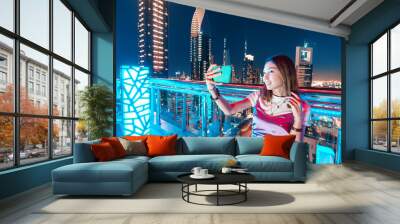 A girl blogger in a rooftop bar takes a selfie photo against the background of a night city with skyscrapers Wall mural