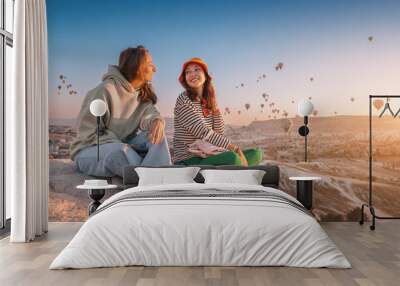 Girl friends sitting on a clifftop viewpoint and admiring view of majestic flying hot air balloons in Cappadocia Wall mural