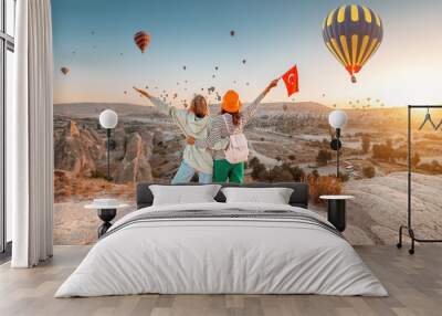 Girl friends travellers with turkish flag, hugging on a viewpoint and admiring view of flying hot air balloons in Cappadocia Wall mural