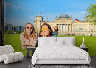 Two Young happy girls wearing sun glasses lying on a grass and have fun in front of the Bundestag building in Berlin. Studying abroad and travel in Germany concept Wall mural