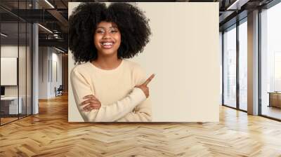 Smiling dark-skinned young woman points with her finger to a space on the right side of the image, isolated on beige background. Wall mural