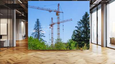 Tower construction cranes and trees against a blue sky with clouds. Boom rotary crane with boom. Construction of apartment buildings in the city. Construction of high-rise buildings. Wall mural