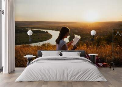 Girl sitting on a hill at sunset and reads a book against the backdrop of a large river. Girl reading book with nice mountain view and river. Wall mural