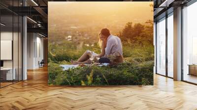 Happy young couple enjoying picnic on the hill. Sunset over hill in the background. Man is hugging and kissing from behind his cute woman. Romance, dating and love concept. Wall mural