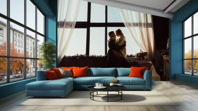 Silhouette of couple near the big window. Gentle embrace and kisses. Beautiful young man and woman is looking at each other and smiling while sitting on windowsill. Stylish interior.  Wall mural