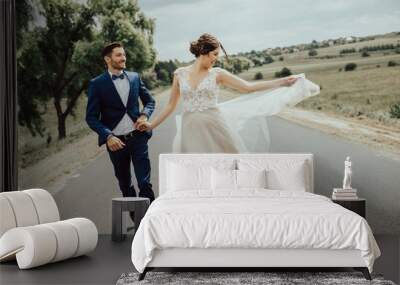 Wedding couple running on the road.Happy young bride and elegant groom running away holding hands.Stylish couple of newlyweds on their wedding day.  Wall mural