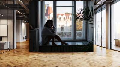 Young thoughtful woman sitting on window sill at home, holding head in hand. Remembering sad moments, feeling lonely. Wall mural