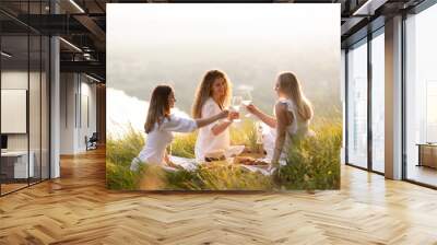 Young women enjoying summer picnic at sunset on the hill. They clinking glasses of wine to celebrate their free time. Wall mural