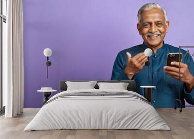 Panoramic wide banner of senior Indian man with blue shirt, grey hair and moustache smiling and holding mobile phone isolated on purple studio background, copy space for text Wall mural